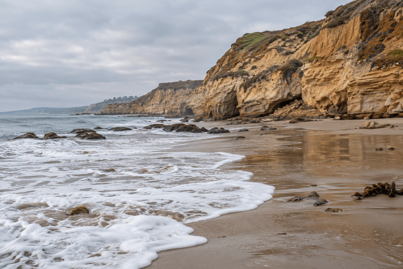 Ocean foam crosses a broad beach beneath Southern California sandstone cliffs.